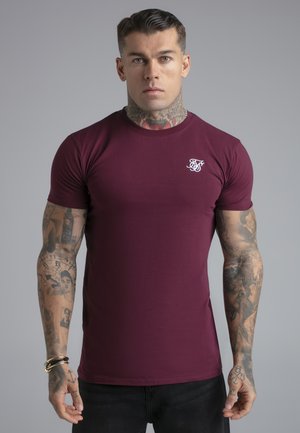 Muskelöser Mann mit Hals- und Arm-Tattoos, der ein eng anliegendes bordeauxrotes T-Shirt und schwarze Hosen trägt, vor einem schlichten grauen Hintergrund stehend.