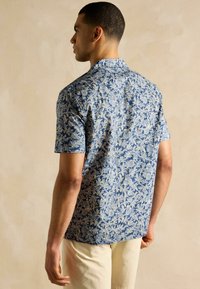 Modrá krátkorpová košeľa s bielym kvetinovým vzorom. Vyrobená z ľahkej tkaniny, vybavená golierom na zapínanie a jemnou textúrou.