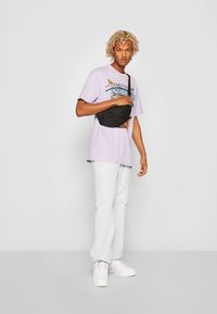 Geantă de umăr neagră, tricou graphic mov cu temă de vară, pantaloni albi și adidași albi cu accente deschise. Păr scurt, creț.