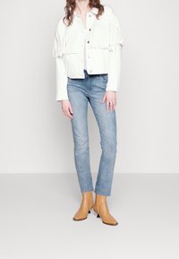 Chaqueta de denim blanca con detalles de flecos, cierre de botones, combinada con jeans de color azul claro y botas de tobillo en tono marrón.