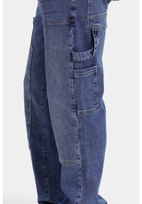Pantalones cargo de mezclilla en un lavado medio, con múltiples bolsillos laterales, corte recto y un sutil desgastado en la tela para darle textura.