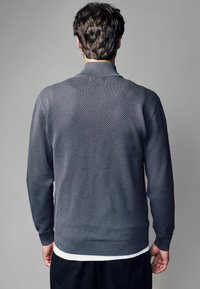 Texturerad, mörkgrå kofta med långa ärmar, ribbade ärmslut och nederkant. Har en hög krage och ett subtilt diamantmönster.