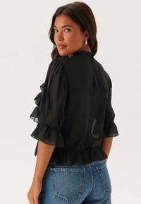 Blusa nera trasparente con maniche a ruffle e orlo peplum, caratterizzata da un collo alto e una texture morbida. Indossata con jeans in denim blu.