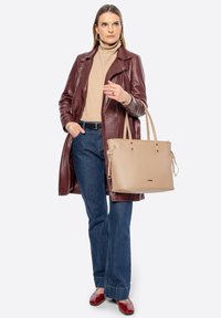 Cappotto di pelle marrone, dolcevita beige, jeans a campana blu. Tiene una grande borsa tote beige con tracolle regolabili. Scarpe rosse con dettagli.
