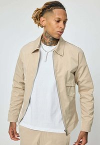 Homme avec des dreadlocks et des tatouages sur le cou portant une veste zippée beige, un t-shirt blanc, un pantalon beige et un collier en chaîne argenté, regardant sur le côté.