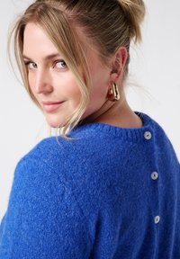 Blauwe wollen trui met een gestructureerde afwerking, ronde halslijn en drie witte knopen aan de achterkant, gecombineerd met gouden oorringen.