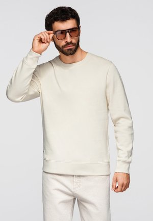 Mężczyzna z ciemnymi włosami i brodą, ubrany w beżową bluzę i jasne spodnie, regulujący brązowe okulary przeciwsłoneczne prawą ręką.