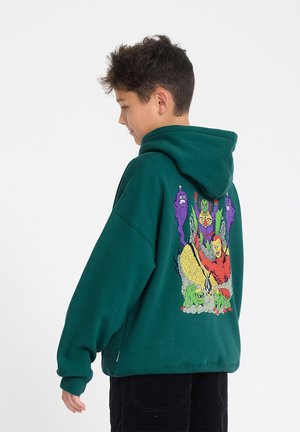 Jongen draagt een donkergroene hoodie met kleurrijke cartoon superhelden en monsters op de achterkant, tegen een effen witte achtergrond.