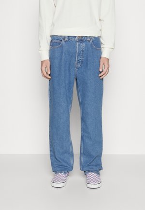 Person trägt weite blaue Jeans, weißes Langarmshirt und lila-weiß kariert Slip-on-Schuhe und steht vor einem einfarbigen Hintergrund.