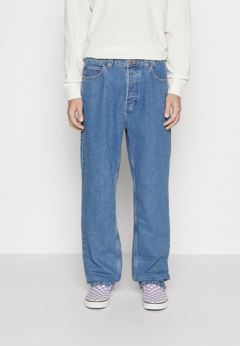 Person trägt weite blaue Jeans, weißes Langarmshirt und lila-weiß kariert Slip-on-Schuhe und steht vor einem einfarbigen Hintergrund.