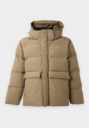 Homme portant une doudoune à capuche beige, pantalon gris foncé, chemise noire et baskets marron, debout avec une main dans la poche de sa veste.