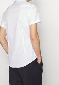 Muž v bílém tričku s krátkým rukávem a černých kalhotách, stojící s jednou rukou v kapse a obrácený zády k jednobarevnému pozadí.