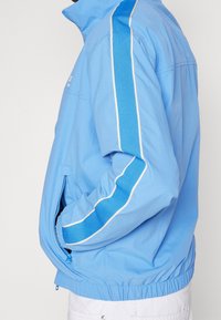 Chaqueta azul claro con cuello alto, que incluye un bolsillo lateral con cremallera, detalles en blanco y rayas verticales texturizadas en las mangas.