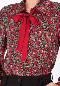 Blouse florale noire, ornée d'un motif de fleurs rouges et de feuilles vertes. Elle possède un grand nœud rouge au col et des bordures en dentelle délicate.