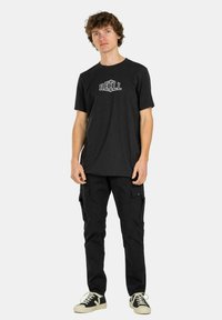 Schwarzes Baumwoll-T-Shirt mit "REELL" Logo, kombiniert mit schwarzen Cargo-Hosen mit Seitentaschen und cremefarbenen Sneakers.