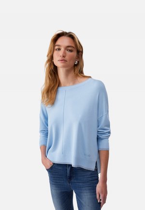 Donna con lunghi capelli biondi che indossa un maglione a maniche lunghe azzurro chiaro e jeans blu, in piedi con una mano in tasca su sfondo bianco.
