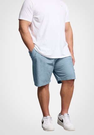 Mann trägt weißes T-Shirt, hellblaue Shorts und weiße Nike-Sneaker, steht mit einer Hand in der Tasche vor einfarbigem Hintergrund.