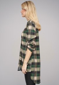 Camicia a quadri verde e beige con maniche arrotolate, chiusura con bottoni e dorso più lungo. Materiale in cotone, texture morbida e vestibilità rilassata.