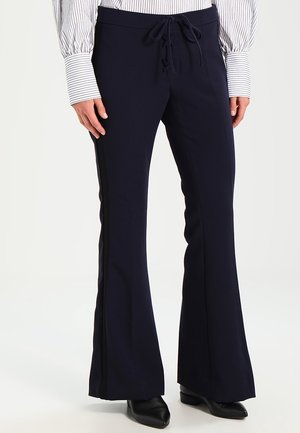 Persona vistiendo pantalones anchos de pierna en color azul marino con cintura de cordón y zapatos negros de punta puntiaguda, combinado con una camisa de manga larga blanca con rayas.