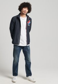 Chaqueta ligera de la marina con cierre de cremallera, logo gráfico rojo y azul, llevada sobre una camiseta blanca, combinada con jeans azules y zapatillas blancas.