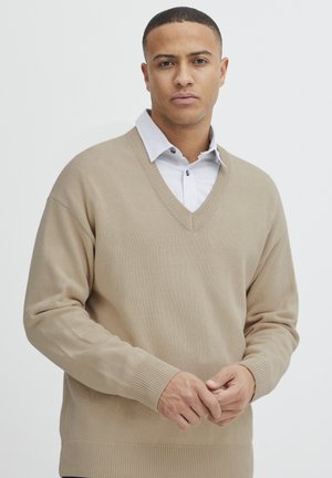Beiger, gestrickter Pullover mit V-Ausschnitt. Verfügt über lange Ärmel sowie gerippte Bündchen und Saum. Getragen über einem hellblauen, gestreiften Hemd mit Kragen.