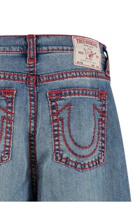 Tył niebieskich dżinsów z czerwonymi szwami, dwie tylne kieszenie oraz biała metka marki True Religion na pasku.