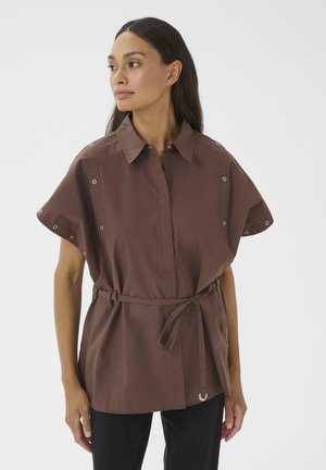 Frau trägt ein lockeres, braunes, kurzärmeliges Hemd mit Gürtel und Metallösen, kombiniert mit schwarzen Hosen, steht vor einem weißen Hintergrund.