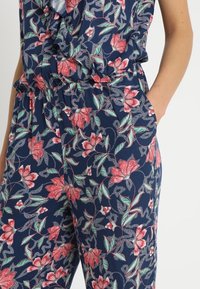 Blommig jumpsuit med en marinblå botten, prydd med rosa och gröna blommönster. Elastisk midja och sidofickor för extra funktionalitet.