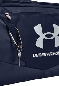 Tamsiai mėlynas "Under Armour" sportinis krepšys su dideliu sidabriniu logotipu, tvirtu užtrauktuku ir raktų pakabuku, pritvirtintu karabineriu.