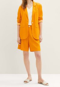 Oranžový lněný blejzr se shodnými kraťasy; bílá košile s V výstřihem pod tím. Model má na sobě béžové skloubené sandály; viditelné knoflíky a kapsy.