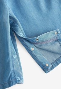 Lys blå denimstof med en glat overflade, der har trykknapper i to nuancer og kontrasterende syninger langs kanterne.