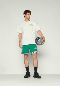 Valge T-särk mitmevärvilise Nike logo, rohelised korvpallishortsid valgete detailide ja logoga, mustad spordijalatsid, hoides korvpalli.