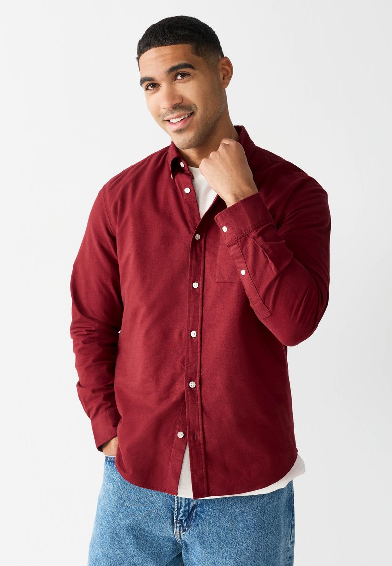 Chemise rouge à boutons avec une poche, en tissu doux, portée par-dessus un t-shirt blanc. Elle comporte des manches longues et des boutons blancs.