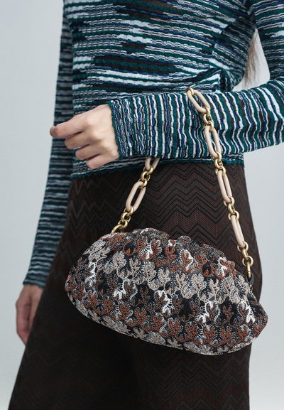 Pessoa segurando uma clutch com padrão floral texturizado e alça em corrente dourada e bege, vestindo uma blusa azul listada e uma saia marrom em padrão zigue-zague.