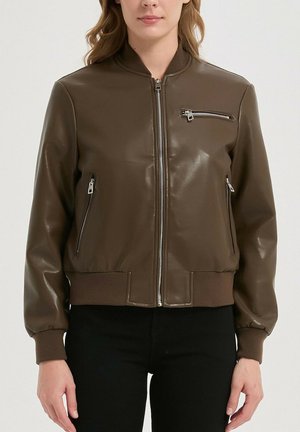 Femme portant une veste aviateur en cuir marron avec des poches zippées et des poignets et ourlets côtelés, associée à un pantalon noir.