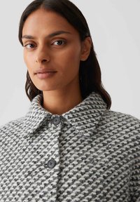 Frau mit mittlerem Hautton und dunklem Haar, die einen strukturierten knopfgeknöpften Mantel in Schwarz und Weiß mit Kragen vor einem einfarbigen Hintergrund trägt.