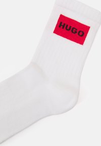 Bijela pamučna čarapa s crvenim pravokutnim aplikacijom s riječju "HUGO" u crnoj boji, rebrastom teksturom i dizajnom do gležnja.