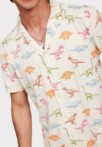 Korte mouw knoopshirt in crème met kleurrijke dinosaurustransformaties. Bevat roze, blauwe, groene en oranje dinosaurussen op een lichte achtergrond.