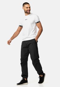 Camiseta de algodón blanca con texto negro en el pecho y rayas negras en las mangas, combinada con pantalones cargo negros y zapatillas negras.