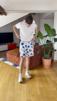 Mężczyzna w białej koszulce, w wzorzystych szortach i trampkach stoi w salonie z brązową kanapą i dużą doniczką z rośliną.