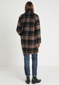 Ternet frakke i brune, sorte og grønne nuancer; lavet af tekstureret stof; midilengde med lige snit; båret over blå jeans og sorte støvler.