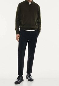 Pull vert foncé à col zippé associé à un pantalon noir et des chaussures noires. Design simple, texture lisse et coupe ajustée.