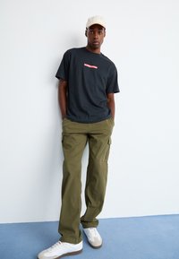 Black t-shirt with red logo, olive cargo pants with side pockets, white sneakers, light beige cap. Model standing against a white wall.
