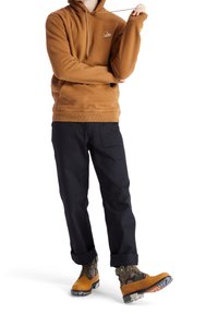 Persona con sudadera marrón, pantalones negros enrollados y botas de senderismo de camuflaje color beige, de pie sobre un fondo blanco.