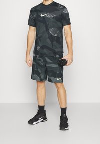 Nike camouflage träningskläder som består av en kortärmad svart och mörkgröna mönstrade skjorta och matchande shorts, med en guldig logotypaccent.