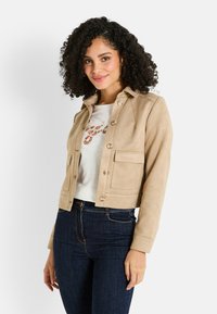 Veste courte en suede beige clair avec poches avant et fermeture à boutons, assortie avec une chemise blanche et un jean bleu foncé.