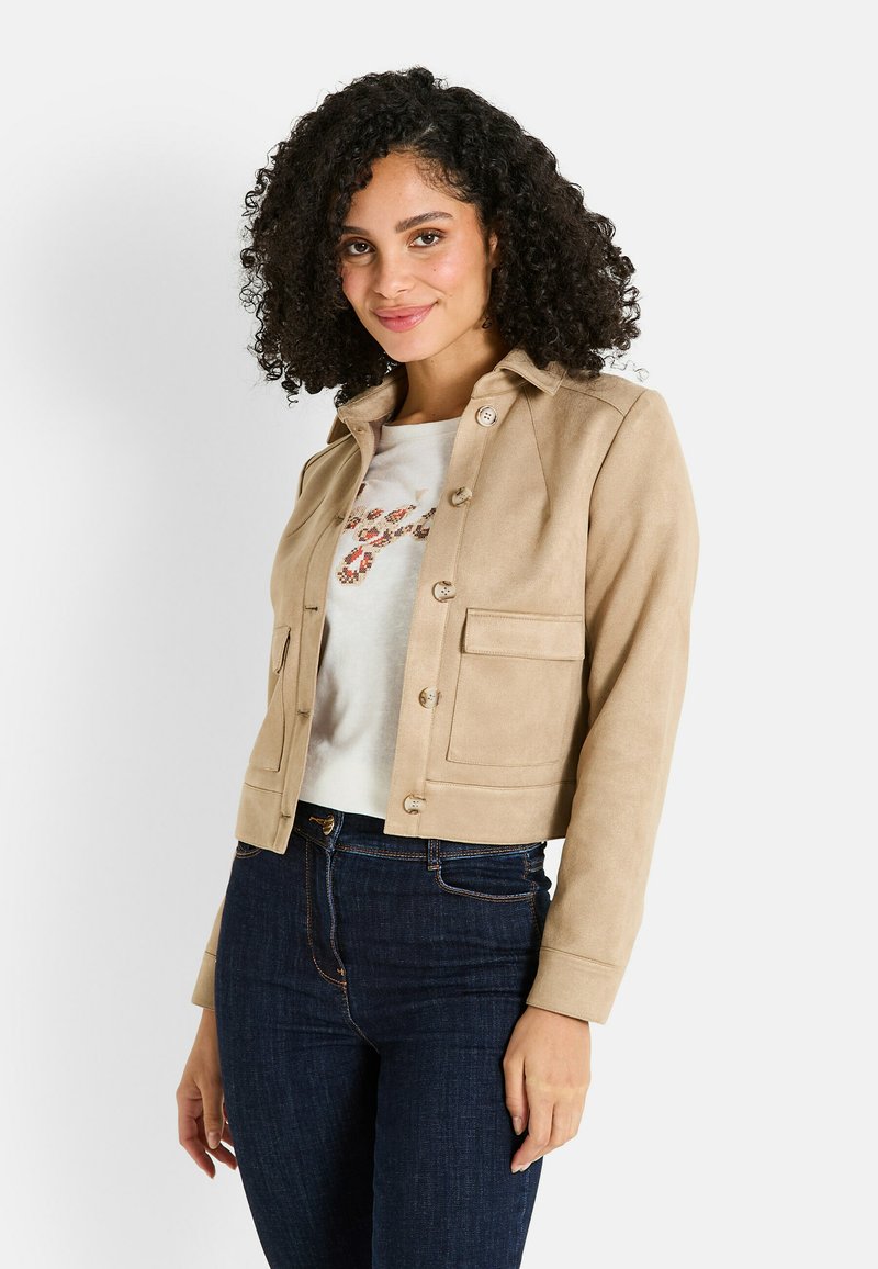 Veste courte en suede beige clair avec poches avant et fermeture à boutons, assortie avec une chemise blanche et un jean bleu foncé.