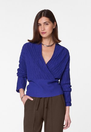 Pull en tricot bleu riche avec un motif texturé, col en V profond et poignets côtelés. Associé à un pantalon marron plissé.