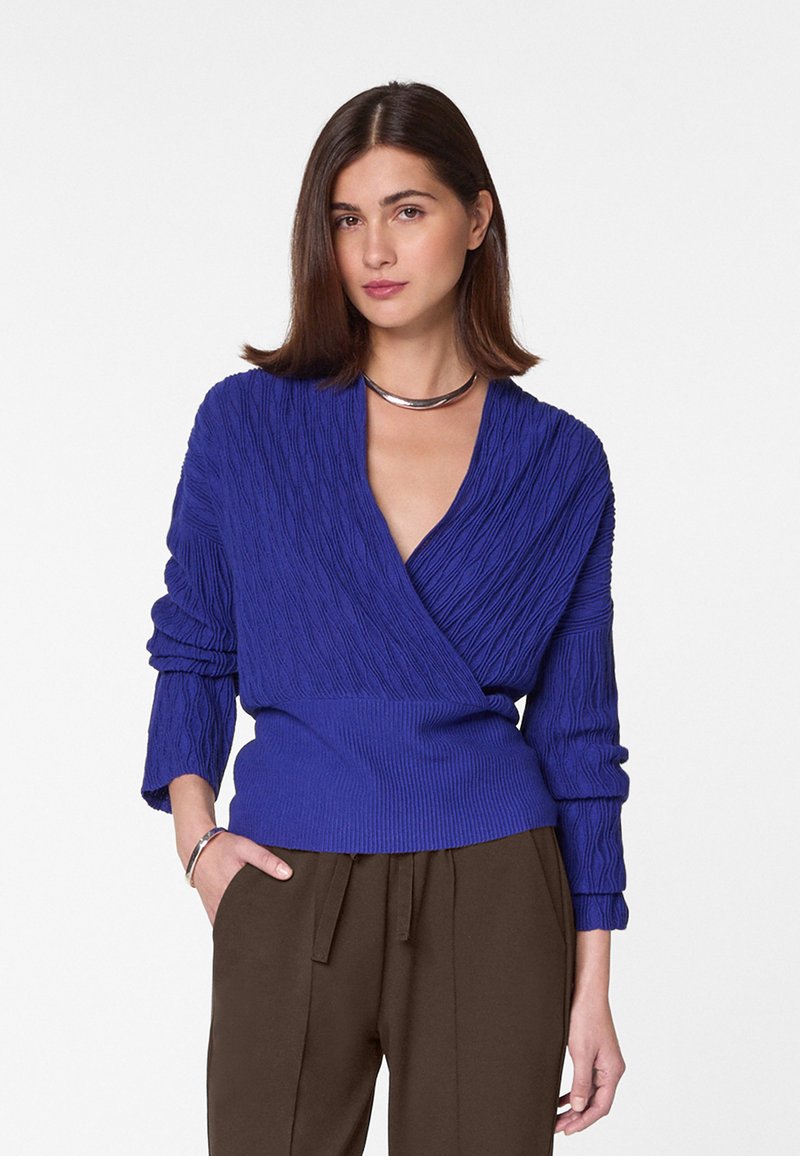 Strickpullover in sattem Blau mit strukturiertem Muster, tiefem V-Ausschnitt und gerippten Bündchen. Kombiniert mit braunen, gefalteten Hosen.