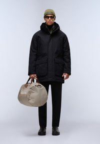 Zwarte gewatteerde parka met twee voorzakken, gedragen over een zwarte coltrui, gecombineerd met zwarte broeken en zwarte schoenen. Houdt een beige sporttas vast.